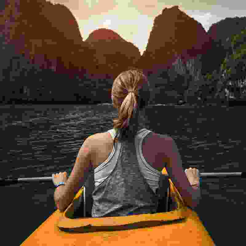 A single woman kayaking on a peaceful river with large rock formations nearby.