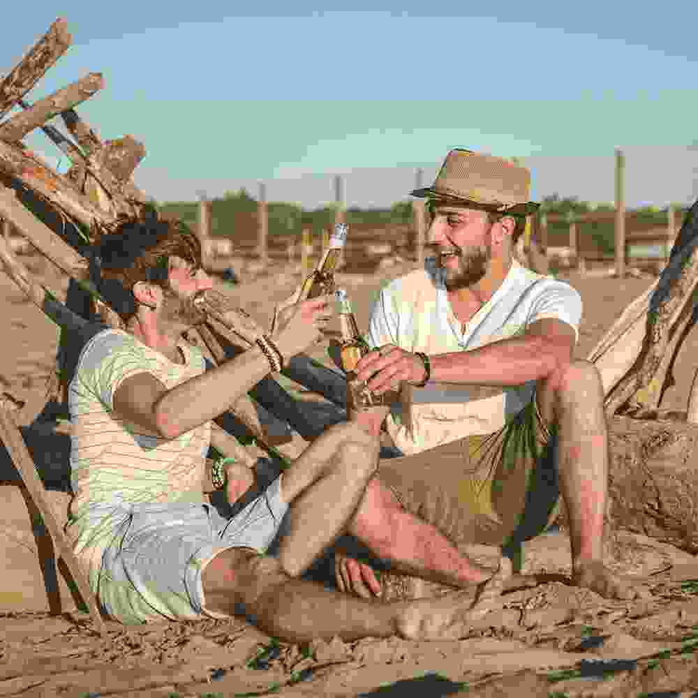 Two dudes chilling on the sandy beach with washed up trees, drinking beers.