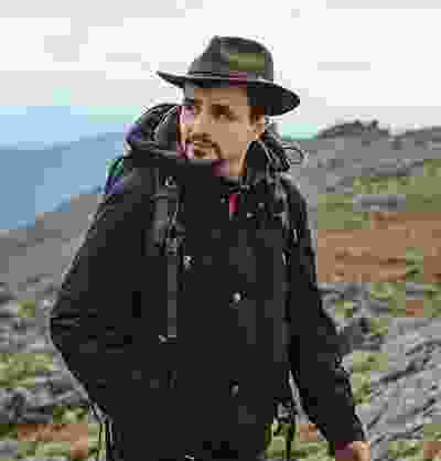 A nonchalant Western man in a fedora hat hiking judiciously in a mountainous area.